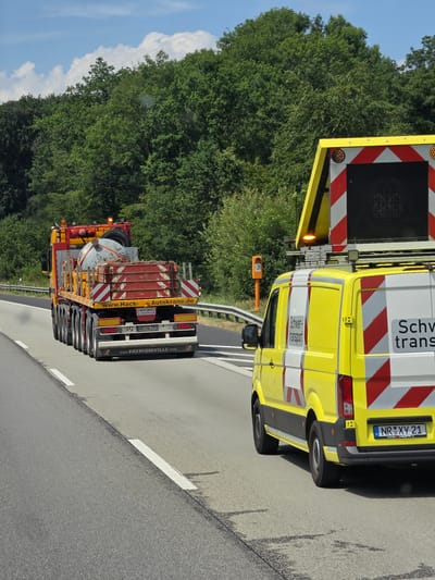 A yellow truck with a large, heavy load is driving on a highway. The load appears to be a large, cylindrical object. The truck has multiple axles and is branded with "www.Hack-Autokrane.de" and "www.FAYMONVILLE.COM". A yellow van with reflective markings and the text "Schwer-transport" is also visible, possibly accompanying the truck. The van has a license plate that reads "NR XV 21". In the background, there are trees and a blue sky with clouds. The setting is a highway with a guardrail and lane markings. The presence of a speed limit sign in orange in the background indicates this is a controlled area. The image appears to be taken in Sessenhausen, Germany, as indicated by the presence of "Sessenhausen" in the data for similar images.
