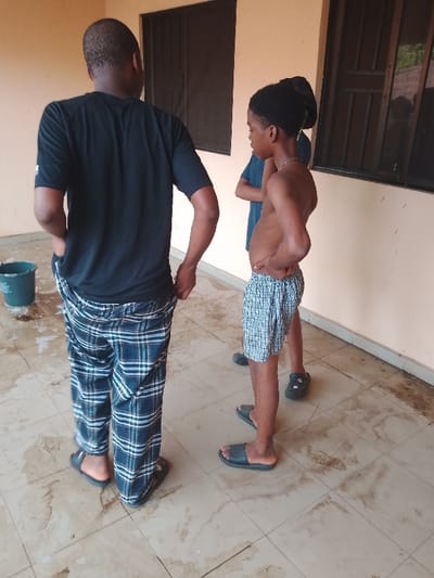 Three individuals stand on a tiled floor in Ufuma, Nigeria, within a light peach-colored structure featuring two dark-framed windows in the background. The light-colored tiled floor displays numerous dark, irregular wet stains.

On the left, an adult male, viewed from behind, wears a black short-sleeved t-shirt with a white logo on the left bicep and dark blue plaid pants. He wears dark slides, with his hands positioned in his pockets. A green plastic bucket is partially visible behind his left leg.

To his right, a young male stands facing left, shirtless, exhibiting a noticeable abdominal protrusion. He wears light blue plaid shorts, dark slides, and has his right hand on his hip. A thin silver-colored chain is around his neck, and a green clip is visible in his dark hair.

Partially obscured behind the shirtless young male is a third individual, wearing a dark blue t-shirt and dark shoes with a star pattern. All individuals are standing in a relaxed posture.