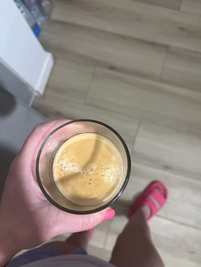 The image depicts an overhead, first-person perspective of an individual's lower torso and extremities in an indoor setting. A left hand, featuring bright pink nail polish on the ring and pinky fingers, holds a clear, cylindrical glass. The glass contains a light brown liquid topped with a significant layer of frothy crema. Below the hand, a fragment of light purple fabric is visible, consistent with clothing. To the lower right, a right foot is visible, clad in a bright pink slide sandal with two distinct straps. The floor is covered with light-toned, plank-style laminate or wood flooring, exhibiting subtle grain patterns. In the upper left quadrant, a section of a white, rectangular cabinet or piece of furniture is present, behind which blurry items, including a plastic bottle and a blue object, are partially visible. The scene is illuminated by even, diffuse lighting.
