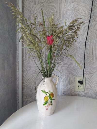 A bouquet of dried grasses and a single red flower is displayed in a light-colored ceramic vase with a lemon and flower motif on the front. The vase has a handle on the right side. The bouquet is tall and bushy, with various shades of green and brown. The vase sits on a white, glossy surface, possibly a table. In the background, a light gray wall is visible with a textured wallpaper featuring a pattern of large, stylized leaves. To the left of the bouquet, a portion of a white pipe or radiator is visible against a gray textured wall. To the right of the bouquet, a black electrical cord hangs down from the top of the frame and plugs into a wall socket. The image appears to be an indoor still life, likely a domestic setting. The image was taken in the city of Gusev, Russia.