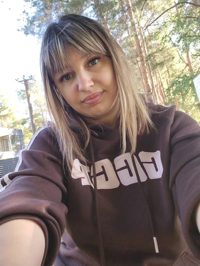 In this close-up, selfie-style photograph, a woman with blonde hair, bangs, and light blue eyes is captured. She is wearing a brown hooded sweatshirt with white and pink lettering on the front. Her lips are slightly parted, and she appears to be smiling subtly. The background suggests an outdoor setting, possibly a park or wooded area, with tall trees and sunlight filtering through the branches. There is a building with a modern architectural style visible on the left side of the frame, and a portion of a white sign can be seen on the far right. The overall lighting is bright, indicating it is daytime. The image does not provide enough context to determine the specific location within Stroitel', Russia.