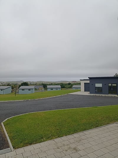 The image depicts a modern residential area with several low-rise structures set against a backdrop of rolling hills under a cloudy sky. In the foreground, a paved driveway curves around a well-maintained green lawn. To the right, a dark-colored, rectangular building with large windows and a glass-paneled door is visible. Further in the middle ground, several light blue, single-story cabins with dark roofs and small windows are spaced out across the grassy landscape. In the background, a cluster of houses and buildings are nestled amongst the green terrain, with distant hills forming the horizon. The overall impression is one of a contemporary, possibly holiday, settlement in a rural or semi-rural setting.