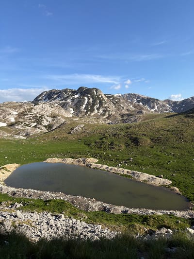 The image depicts a mountainous landscape in Boričje, Montenegro. A long, dark lake is situated along a grassy hillside that ascends towards a rugged, rocky mountain. Patches of snow remain on the mountain's rocky slopes. The sky is blue with scattered wisps of clouds. The vegetation appears lush in areas, contrasting with the barren rocky terrain. No people are visible.