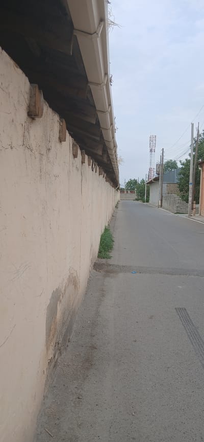 The image depicts an outdoor scene, likely in Andijani, Uzbekistan, on a cloudy day. The perspective is low and angled, looking up a paved walkway that slopes gently upwards. To the left of the walkway, a tan-colored gutter system runs along a textured wall. The gutter appears to be made of metal and is supported by wooden beams. Small, round tree stumps are visible along the walkway's edge.
Beyond the walkway and to the right, a paved road is visible. There are tire marks on the road, suggesting recent vehicle traffic. In the distance, along the road, there are buildings, trees, and what appear to be communication towers, one of which is red and white. Electrical poles and wires are also present, adding to the sense of a developed area. Green grass grows in patches along the base of the wall and on the side of the road. The overall atmosphere is one of an ordinary day in a semi-urban or town setting. There is no text visible in the image.