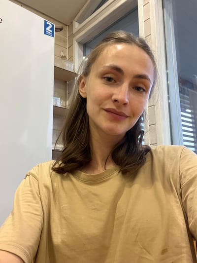 The image is a selfie of a young woman with light brown hair, looking directly at the camera. She is wearing a light brown t-shirt. Her face is partially illuminated, with shadows indicating a light source to her right. The background appears to be an indoor setting with wooden paneling on the walls and a window with horizontal blinds. To the left of the woman, a white refrigerator is partially visible, with a blue and white sticker that reads "2 ГОДА ГАРАНТИИ" (2 YEARS WARRANTY) in Russian. The overall impression is a casual, personal snapshot taken in a home environment, possibly in Krugloye, Russia.
