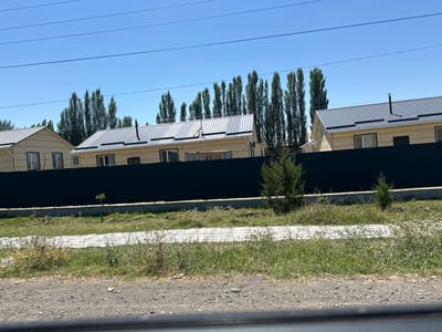 In the city of Тынчтык, Kyrgyzstan, a row of modern, cream-colored houses with dark metal roofs stands behind a dark, solid fence. The houses have white trim around their windows and doors, and some appear to have air conditioning units. Tall, slender trees line the street behind the houses, creating a vertical contrast to the horizontal lines of the architecture. In the foreground, a patch of dry grass and weeds borders a paved walkway. Power lines are visible against the clear blue sky.
