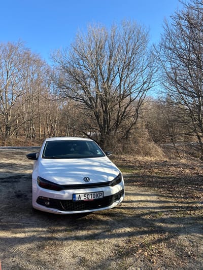 A white two-door Volkswagen Scirocco is parked on an unpaved, dirt and gravel surface with scattered dry leaves and sparse dry vegetation. The vehicle faces the viewer, angled slightly left, and prominently displays a Bulgarian license plate "A 5010 KP" on its front bumper. The headlights feature a dark tint or cover. An object hangs from the interior rearview mirror. Long shadows from unseen objects extend across the ground from the right. The background consists of a dense stand of leafless deciduous trees under a clear blue sky, indicative of a late autumn or winter season. No individuals are present in the scene. The location is Ichera, Bulgaria.