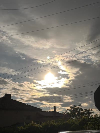 The image shows a dramatic sky over Aidemir, Bulgaria, with the sun breaking through dark, dramatic clouds. A sun dog is visible, a bright optical phenomenon caused by the refraction of sunlight by ice crystals in the atmosphere. The sun itself is partially obscured by clouds, creating a halo effect. In the foreground, the roofs of houses and dense foliage are visible. Electrical wires crisscross the sky, adding a man-made element to the natural scene. The overall impression is one of a dynamic and somewhat foreboding sky, with hints of beauty in the sun dog.
