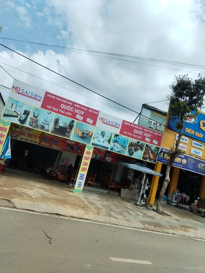The scene is a street-level view of two adjacent stores in Pờ Y, Vietnam. The store on the left has a large sign reading "HD SAISON" and "QUỐC HUY," advertising appliances, phones, and furniture with installment plans. Images of furniture and appliances are displayed on the sign. Inside the store, wooden furniture is visible. A smaller sign in front advertises promotions, specifically a lucky draw (bốc thăm trúng thưởng), and mentions "Nội thất cao cấp" (high-class furniture). A small snake is visible on the road. The adjacent store on the right, painted yellow and blue, advertises electronics brands such as Sony, Sharp, Samsung, and Aqua. A tree stands in front of this store. A motorcycle is parked near the second store. The sky is cloudy.