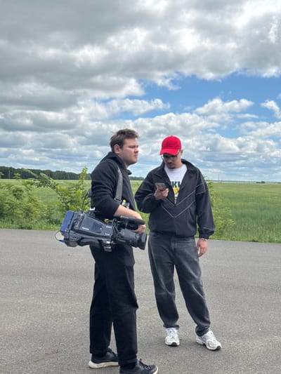 Two men are standing on an asphalt road near a green field under a cloudy sky. The man on the left is wearing a black hoodie and black pants. He is carrying a professional video camera on his shoulder. The man on the right is wearing a black jacket with white stripes, a white t-shirt, dark jeans, and a red baseball cap. He is looking at his phone, which he holds in his right hand. The background consists of a large green field and a line of trees in the distance. The sky is filled with large, cumulus clouds, with patches of blue sky visible. The setting appears to be a rural or semi-rural area.