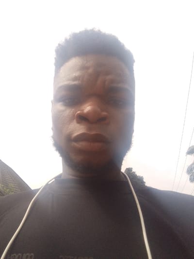 The image displays a close-up, upward-angled selfie of a young adult male, situated outdoors in Agukwu Nri, Nigeria. The individual, of apparent African descent, has short, dark, textured hair and dark facial hair along his jawline and chin. He is wearing a dark, possibly black, crew-neck shirt. A white cable, consistent with an earbud cord, is draped around his neck, extending over his right shoulder. His gaze is directed towards the camera, and his forehead exhibits slight furrows. The background is predominantly an overexposed bright white or light gray, indicative of an overcast sky or bright ambient light. To the viewer's left, a dark-colored tiled roof structure is partially visible. To the viewer's right, dark green foliage, consistent with trees or palm fronds, and thin dark lines, likely overhead utility cables, are discernible against the bright background. The precise location within Agukwu Nri, Nigeria, is not identifiable from the visual evidence presented.