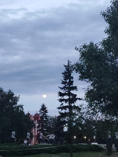 The image depicts an outdoor scene at dusk in Slavgorod, Russia. The sky is overcast with heavy, gray clouds, creating a subdued atmosphere. A full moon is visible in the sky, appearing as a bright, luminous orb peeking through the clouds. In the foreground, several busts of people are displayed on dark pedestals, interspersed with lush green bushes and manicured hedges. Tall trees, including a prominent evergreen, flank the busts and the building in the background. The building itself is a striking structure with red and white accents and distinctive turret-like rooftops, suggesting it might be a historical or governmental building. The lighting is dim, indicative of the twilight hours, with a few streetlights also visible in the distance. The overall mood is serene and perhaps a little melancholic due to the twilight setting and the static nature of the busts. There is no visible text in the image.