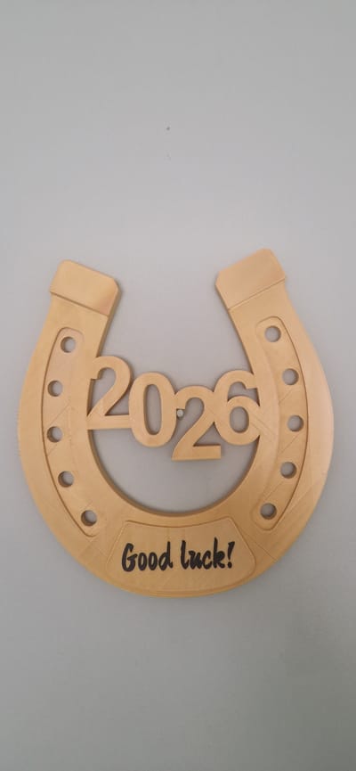 A light brown, wooden horseshoe ornament is displayed against a plain, light gray background. The ornament is positioned vertically, with the open end pointing upwards. The numbers "2026" are carved into the center of the horseshoe, and below this, the words "Good luck!" are written in a dark, cursive font. The horseshoe has decorative circular indentations on each side. There are no people or any other distinguishing environmental features visible. The location of this image is unknown and cannot be determined as South Holland, United Kingdom, or any other specific city.