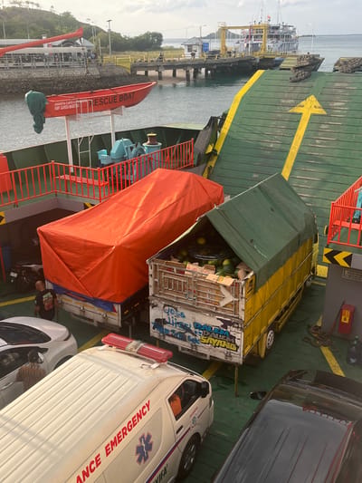 In Upt Tambak Sari, Indonesia, a ferry is docked at a port. On the ferry's deck, a red rescue boat is suspended by a crane. Several vehicles are parked on the ferry, including an ambulance with "AMBULANCE EMERGENCY" written on its side, a white car, a yellow truck with a green tarp covering its cargo of watermelons, and a truck covered in an orange tarp. A person wearing a black t-shirt is standing next to the orange-covered truck, and another person is visible inside the white car. The ferry deck is painted green with yellow directional arrows. In the background, a large ferry is docked at the port, and a bridge extends into the water. The sky is overcast.