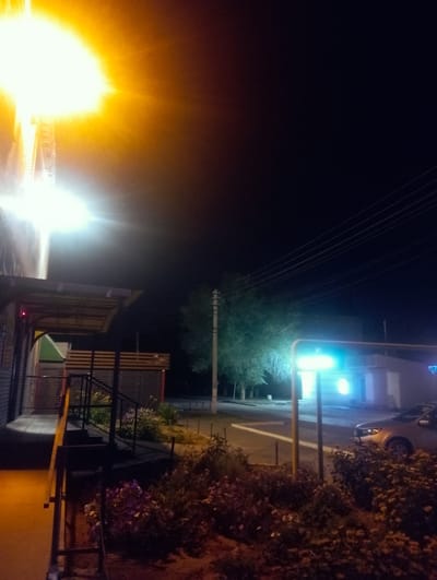 The image depicts a nighttime scene on a street in Znamensk, Russia. A bright, overhead light illuminates the left side of the frame, casting long shadows. In the foreground, a sidewalk leads to a building with a covered entrance and a set of stairs. To the right of the stairs, a garden area with various plants is visible. Further down the street, a car is parked near a building with a bright, blue-green light. A utility pole with power lines runs diagonally across the upper right portion of the image. The overall atmosphere is dimly lit, with artificial lights dominating the scene.