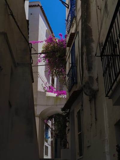 The photograph captures a narrow alleyway in Sperlonga, Italy, with whitewashed buildings on either side. Vibrant pink bougainvillea cascades down from a balcony on the right, contrasting with the weathered white walls of the buildings and the deep blue sky above. A wrought-iron balcony railing, painted a dark blue, is visible on the upper part of the right building. The sunlight casts sharp shadows, indicating a bright, sunny day. The overall scene evokes a sense of Mediterranean charm and timelessness.