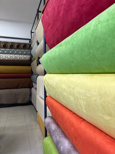 The image depicts a retail setting, likely a fabric store, displaying a variety of fabric rolls stacked on shelves. The rolls are oriented horizontally, with their ends facing the viewer. Prominent colors include red, bright green, pale yellow, and orange, with other neutral tones and patterned fabrics visible in the background. The fabrics appear to be soft and possibly velvety or plush in texture. The shelving units are metal, suggesting a commercial display. The floor is tiled with large, light-colored square tiles. The lighting appears to be overhead and artificial, creating a well-lit environment. The context of La Victoria, Peru, suggests this is a business located within that district, known for its commercial activity, particularly in textiles and apparel.
