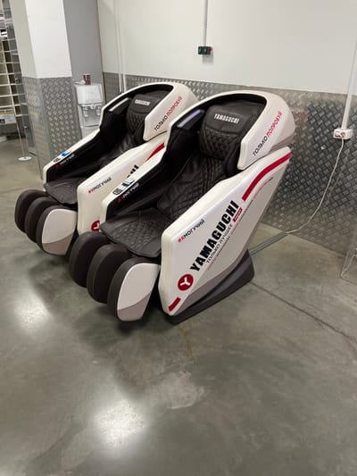 Three high-tech Yamaguchi brand massage chairs are arranged in a row on a light gray concrete floor within an indoor commercial or public space in Sholokhovo, Russia. The chairs are predominantly white with dark brown or black quilted seating surfaces and red accent stripes. Each chair features the "YAMAGUCHI" logo on the headrest and side panels. Text in Cyrillic is visible on the chairs: "ТОЛЬКО ПОПРОБУЙ" (JUST TRY IT) on the upper sections, and "#ЯМОГУЧИЙ" (#IMIGHTY) on the armrests, along with "ТОЛЬКО ЛУЧШЕЕ" (ONLY THE BEST) and "УКРЕПЛЯЕТ СТОЙКИЙ НАДЕЖНЫЙ КОСТРОПОЛЬ" (STRENGTHENS A STABLE RELIABLE KOSTROPOL') on the lower side panels of the rightmost chair, which also displays "AIRPORT" below "YAMAGUCHI". A small digital display is present on the armrests of the two leftmost chairs.
The background wall features a lower section covered in silver diamond-plate metal sheeting and an upper section of plain white wall. A white floor-standing water cooler with a brown paper cup resting on top is positioned on the left, next to a vertical white column that is also partially covered with diamond plate. An electrical outlet with red and green buttons is mounted on the white wall above the diamond plate, and a white power strip is affixed to the diamond plate further to the right. White shelving units are partially visible behind the column on the far left. No individuals are present in the scene.