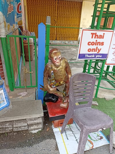 Outside a building with a yellow accordion-style gate in Ban Hin Sam Kon, Thailand, sits a metal monkey statue holding airplane replicas, next to a plastic chair and a sign that reads "Thai coins only".