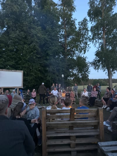 This is a medium shot, taken from a low angle, of an outdoor gathering at dusk in a rural area that could be in Завалочыцкі сельскі Савет, Belarus. A group of approximately 30-40 people, a mix of adults and children, are seated on wooden benches made from pallets around a bonfire. A large, white projection screen is visible on the left side of the frame, suggesting a movie or presentation is planned or in progress. The background consists of trees and a grassy field with a line of trees in the distance. The lighting is dim, with the bonfire providing a warm glow and a few string lights overhead illuminating the scene. The people are casually dressed and appear to be engaged in conversation or watching something. Some individuals are standing, observing the crowd, while others are seated, facing the bonfire or the screen. The overall atmosphere seems relaxed and communal.