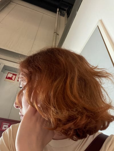 An angled close-up shot depicts a fair-skinned person with reddish-brown hair. The person is wearing glasses and a light-colored shirt. A brown strap is visible on their shoulder. The background includes a white ceiling with a recessed light fixture, a wall, and a fire alarm. The location is Lautzenhausen, Germany.