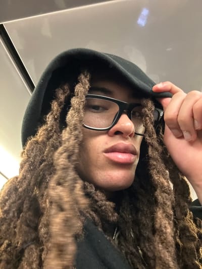 A person wearing glasses and a black hoodie faces the camera. Their hand pulls the hoodie’s top corner. They have brown dreadlocks. The photo appears to be taken indoors with a white background. The focus is primarily on the person's face and hair, with a partial view of their upper body.