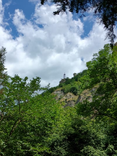 This media file shows a view of a rocky hillside covered in lush green trees and vegetation. At the very top of the hill, a tall metal tower with antennae is visible. The sky above is a mix of bright blue with fluffy white clouds. The foreground is dominated by dense foliage from trees, with branches and leaves partially obscuring the view of the hill and sky. The overall scene suggests an outdoor setting in a natural environment, possibly a mountainous or hilly region, under a partly cloudy sky. The time of day appears to be daytime, with bright light illuminating the scene. There is no visible text in the image. The location context is Madzhalis, Russia.