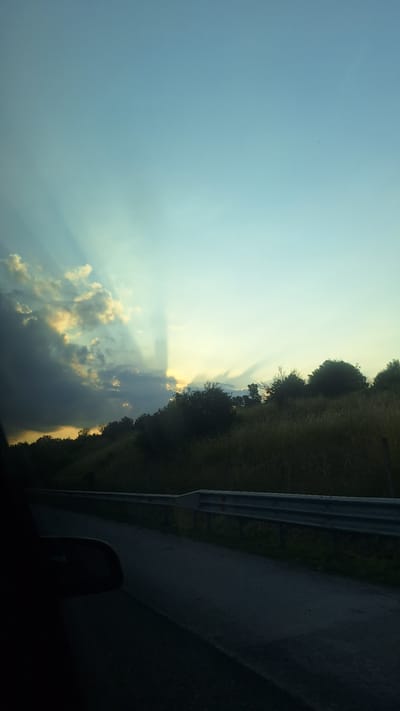 The image depicts a sunset scene viewed from a vehicle moving along a road. The sky is a gradient of pale blue to a lighter hue near the horizon. Sunbeams, also known as crepuscular rays, emanate from behind a band of clouds on the left side of the frame, casting dark shadows across the sky. The clouds themselves are a mix of dark gray and lighter, yellowish tones where illuminated by the sun.
Below the sky, a hilly landscape is visible, covered in dry, tall grass and scattered trees. The vegetation appears dense in some areas and sparser in others. A guardrail runs along the right side of the road, separating it from the grassy embankment. The road itself is asphalt, and appears to be a two-lane highway. The lower left portion of the image is dominated by the interior of the vehicle, showing a dark silhouette that includes what appears to be a side mirror. The lighting suggests either dawn or dusk, with the sun low on the horizon. The scene is set in a natural, rural environment. Based on the provided context, the location is Ravnishte, Bulgaria.