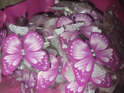 The image displays a close-up of numerous decorative purple butterflies with white accents, each adorned with small pearl-like embellishments. These butterflies are arranged in a bouquet-like fashion, wrapped in transparent plastic and nestled within pink tissue paper. The butterflies appear to be favors or decorations, possibly for an event. The scene is indoors, with artificial lighting casting shadows and reflections on the plastic wrapping. The background is a blur of pink and dark tones, suggesting a box or a larger arrangement. There are no people or identifiable landmarks visible in the image, and therefore no specific location within the city of Hlusk, Belarus can be determined.