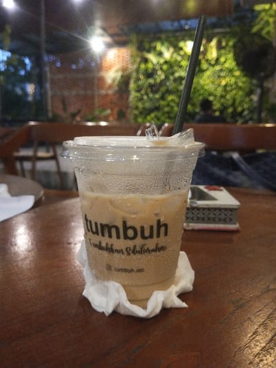 A plastic cup containing an iced coffee beverage is prominently displayed on a dark wooden table in a cafe located in Pontianak, Indonesia. The transparent cup, exhibiting condensation on its exterior, holds a light brown liquid with visible ice cubes and features the brand "tumbuh" with the tagline "tumbuhkan silaturahmi" and an Instagram handle "@tumbuh.idn". A dark straw protrudes from the clear plastic lid. The cup rests on a crumpled white paper napkin. To the immediate left, a portion of another crumpled white napkin is visible. To the right, a decorative ashtray with a white top and a black and white patterned base is present, slightly out of focus. The blurred background reveals a warm-lit interior with several bright overhead lights, a prominent green vertical garden on the right, and a textured brick wall section. Dark wooden tables and chairs are discernible, along with the silhouette of a person seated further in the background.