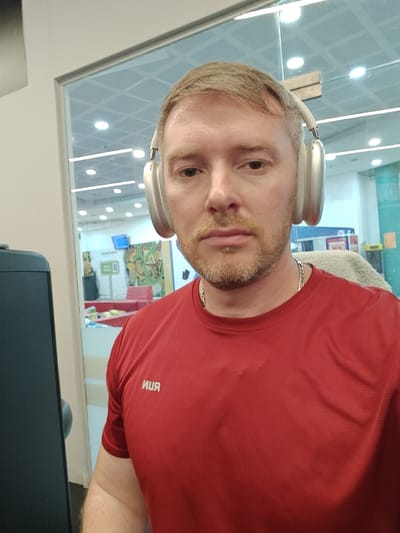 This image depicts an adult male captured in a selfie or close-up portrait, located indoors in the city of Nahariya, Israel. The male subject has light skin, short light brown hair with some visible graying, and a short, light brown beard. He is wearing light-colored, over-ear headphones and a red short-sleeved athletic shirt with the mirrored text 'RUN' in white on the left chest. A silver-colored chain is visible around his neck. He maintains a direct gaze towards the camera. To his left, a dark, vertical object, possibly a monitor or piece of furniture, is partially visible. The background consists of a large glass partition, through which an brightly lit interior space can be seen. This background area features a grid ceiling with multiple recessed light fixtures. Visible beyond the glass are colorful, abstract elements, including a light blue sign, a green sign, orange, green, and yellow shapes, and a red object, possibly a bench or piece of furniture. Reflections of overhead lights are present on the glass surface.