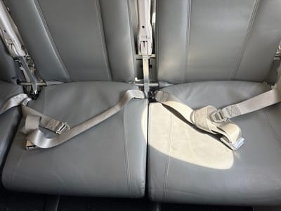 The image displays the interior of a vehicle, consistent with a public transport bus or van, located in Tijuana, Mexico. Two adjacent, empty passenger seats are visible, upholstered in textured gray vinyl or leather. Each seat features a matching gray fabric lap belt with a silver-colored buckle mechanism, both unbuckled and resting on the seat cushions. The metal support frames of the seatbacks are partially exposed between the seats and at the base of the seatbacks. Sunlight illuminates the right seat cushion and the corresponding seatbelt, casting a bright spot and indicating a nearby window to the right of the frame. No individuals or objects other than the vehicle's fixed components are present.
