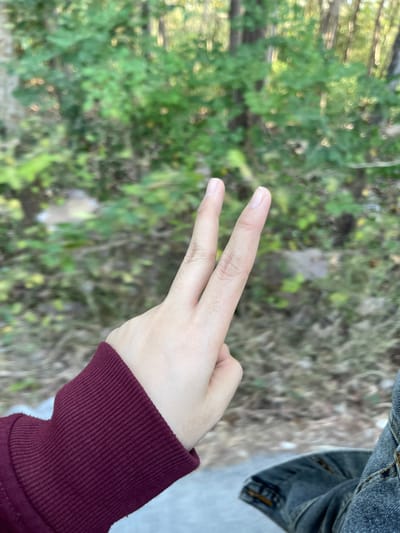 In Seling Kudul, Indonesia, a close-up shot features a person's left hand making a peace sign, with two fingers extended. The hand is clad in a burgundy sweater cuff, and the person is wearing denim jeans. The background is blurred, showing a wooded area with green foliage and trees.