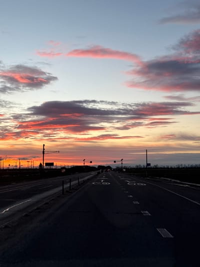 The image displays a wide, multi-lane highway at either sunrise or sunset, characterized by a dramatic and colorful sky. The perspective is looking down the road as it stretches into the distance.

The upper two-thirds of the image are dominated by the sky, filled with long, stretched-out cloud formations. These clouds are painted with vibrant hues of red, orange, and pink, particularly closer to the horizon, which contrasts with darker grey and purple sections within the cloud masses. Higher in the sky, the colors transition to softer yellows and oranges near the horizon, fading into a pale blue and light grey as the sky extends upwards. The lighting suggests a low sun, casting warm tones on the underside of the clouds.

The lower third of the image shows a dark asphalt highway, appearing wide with multiple lanes. Clear white road markings are visible on the surface. Two large, white numbers "60" are prominently painted on the road, indicating a speed limit or lane designation. Additionally, white dashed lines separate the individual lanes.

Along the right side of the highway, a metal guardrail or barrier runs parallel to the road's edge. In the mid-ground, a series of dark silhouettes against the illuminated sky reveal utility poles, streetlights, or gantry structures positioned above or alongside the highway. A faint, dark line on the distant horizon suggests a treeline or landmass. The overall scene evokes a sense of travel or contemplation during a beautiful time of day. No people or vehicles are clearly discernible.