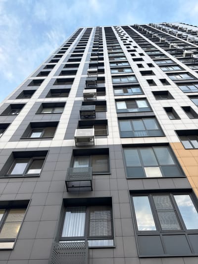 A low-angle, wide shot captures the facade of a modern, tall apartment building under a cloudy sky. The building, with its many windows and sleek, tiled exterior, is angled upwards and to the left, dominating the frame. The facade is characterized by a repeating pattern of white and grey tiles, interspersed with dark-framed windows. Some windows appear to be open, with what look like air conditioning units or ventilation systems visible in the openings, protected by perforated metal screens.
The building's design is contemporary and geometric, with a sense of verticality emphasized by the upward perspective. The sky is a soft blue with scattered white clouds, suggesting a daytime scene, possibly overcast or with the sun behind the clouds. There are no people visible in the image. The overall impression is one of urban architecture, highlighting the structured and modern design of the residential building. The location context indicates the building is in Лесные Поляны, Russia, though no specific landmarks are visible to confirm this. No text is present in the image.