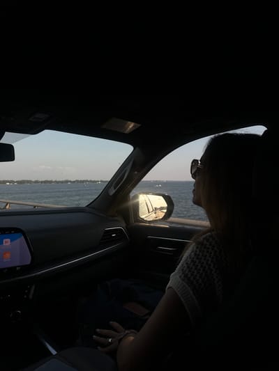 The image depicts the interior of a vehicle from the driver's side perspective, looking towards the passenger and the exterior environment. An adult female is seated in the front passenger seat, silhouetted and facing right, looking out the window. She wears sunglasses, a light-colored knit top, and a ring on her left ring finger. Her left hand rests on her lap. The vehicle's dark dashboard is visible, featuring an infotainment screen displaying "71°" and interface elements. Sunlight reflects prominently off the passenger-side rearview mirror, showing a road surface and sky. Outside the window, a wide body of water, possibly a river, bay, or large lake, is visible under a light blue sky with faint clouds. A distant, tree-lined landmass is discernible across the water, with several small vessels or buoys scattered. A bridge railing or barrier is present between the vehicle and the water. The scene occurs in the city of Montgomery, United States.