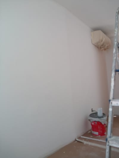 This media file depicts an indoor scene, likely a room undergoing renovation or painting. The dominant subject is a large, pale wall that occupies most of the frame. In the upper right corner, a part of a ladder is visible, suggesting ongoing work. To the left of the ladder, near the ceiling, an air conditioning unit is covered with a light-colored protective material.
On the floor, in the lower right quadrant of the image, a red bucket is placed on a piece of white material, possibly plasterboard or a drop cloth. The bucket is partially covered with a plastic bag, and a clear plastic cup or container is placed on top of it. There is some white debris scattered on the floor around the bucket, indicating painting or plastering activities. The overall impression is one of a work in progress, with tools and materials set up for a renovation project. The lighting suggests it is daytime, as is typical for such work. There is no visible text in the image. The location context of Nueva Andalucía, Spain, suggests a residential or commercial interior in that region.