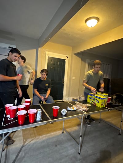 The image depicts an indoor scene in South Bend, United States, featuring four adult males in what appears to be a residential common area. The room is illuminated by an overhead ceiling fixture and shows light-colored walls, a dark-paneled door in the background, and a grey floor.
On the left, a male with dark hair and a dark t-shirt stands, holding a red plastic cup. To his right and slightly behind him, another male with lighter hair and a light t-shirt is walking towards the right, appearing to look at something on his left wrist. In the center, a third male with dark hair and a dark t-shirt is seated on a low stool or chair, engaged with a mobile phone. On the right, a fourth male with short brown hair and an olive-green t-shirt stands behind a black folding table. He is actively interacting with a yellow cardboard box labeled "TWISTED TEA," which contains multiple cans.
The black folding table is central to the foreground. On its left side, seven red plastic cups are arranged in a formation, with one silver aluminum can (possibly "Natty Light") standing upright. On the table's right side, another red plastic cup, a crumpled silver aluminum can, and a clear plastic bag containing snacks are visible next to the "TWISTED TEA" box. The arrangement of cups suggests a recreational activity.