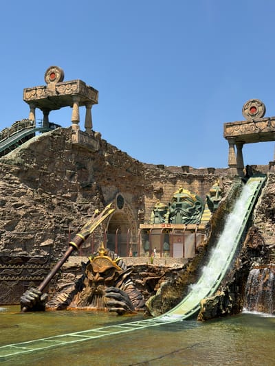 The image shows a water ride attraction at a theme park in Castelnuovo del Garda, Italy. The ride features a steep water slide descending from a rocky, artificial mountain structure. At the base of the slide, a large, bronze statue of a mythical creature emerges from the water, holding a spear. The structure is decorated with architectural elements resembling ancient Egyptian or fantasy themes, including large sculpted heads and decorative columns. A roller coaster track is visible on the left side of the image, suggesting it is part of the same attraction or a nearby one. The sky is clear and blue. A few people are visible in the background near the base of the attraction.