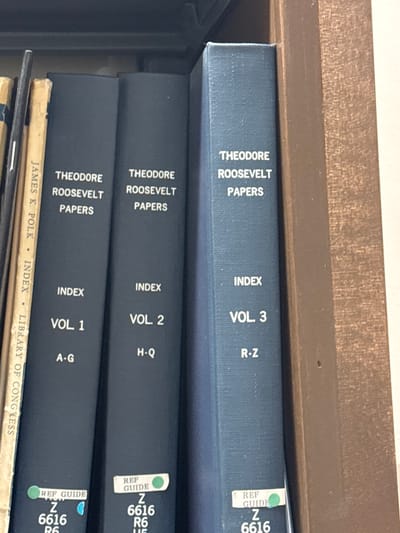 The image displays a section of a wooden bookshelf containing several historical reference volumes, located within an indoor setting in the Town of Vestal, United States. Three prominent dark blue, cloth-bound books are visible, all titled "THEODORE ROOSEVELT PAPERS" and subsequently "INDEX." These are individually labeled as "VOL 1 A-G," "VOL 2 H-Q," and "VOL 3 R-Z" from left to right. Each of these volumes has a white "REF GUIDE" label near the bottom of its spine, marked with "Z 6616" and partially covered by a circular green sticker. To the far left, a tan-colored book titled "JAMES K. POLK INDEX LIBRARY OF CONGRESS" is partially visible. A thin, dark object is situated between the Polk volume and the first Roosevelt volume. The books are arranged vertically on a wooden shelf, with the right side of a brown wooden vertical shelf support visible. No individuals are present, and no actions are occurring.