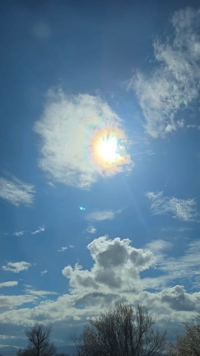 The image shows a bright sun surrounded by a colorful halo in a sky filled with scattered white clouds. Some tree branches are visible on the right side of the image. This scene is most likely in the city of Slatino, Bulgaria, based on the prompt.