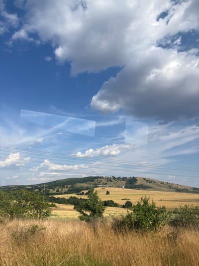 The image depicts a scenic rural landscape under a bright blue sky dotted with white and grey clouds. The foreground is dominated by tall, dry grass, suggesting a summer or early autumn setting. In the midground, a field of golden crops, likely wheat or barley, stretches towards a gently sloping hill. The hill is covered with sparse vegetation, including scattered trees and some patches of green. Several power lines are visible spanning across the scene, supported by utility poles. Two rectangular reflections, possibly from a window, are superimposed on the sky and the upper portion of the landscape. The overall impression is one of tranquility and vastness. The location in Smilets, Bulgaria, is not explicitly depicted by any recognizable landmarks or specific geographical features within the image itself, but the landscape is consistent with the general rural environment of such an area.
