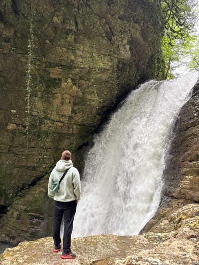 The image depicts a person standing near a waterfall. The person is wearing a light-colored hoodie, dark pants, and red shoes. They are facing towards the waterfall, hands on their hips, with a small bag slung across their shoulder. The waterfall is situated within a rocky cliffside, with green foliage visible at the top. The rock formations surrounding the waterfall are textured and weathered. The location is Tsabal, Abkhazia.
