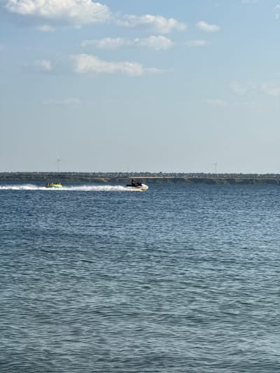 In Ukrainka, Ukraine, on a clear day, two personal watercraft are navigating a wide body of blue water. The craft on the right is a jet ski, ridden by a single person wearing dark clothing, who is kicking up a significant amount of white spray as they move. To the left, a yellow inflatable tube, possibly for recreational towing, is being pulled by an unseen force, creating a smaller wake. Several individuals are visible sitting on the tube. In the background, a low, green-topped embankment runs along the horizon, dotted with scattered houses and a prominent wind turbine. The sky above is a pale blue, with a few wispy white clouds.