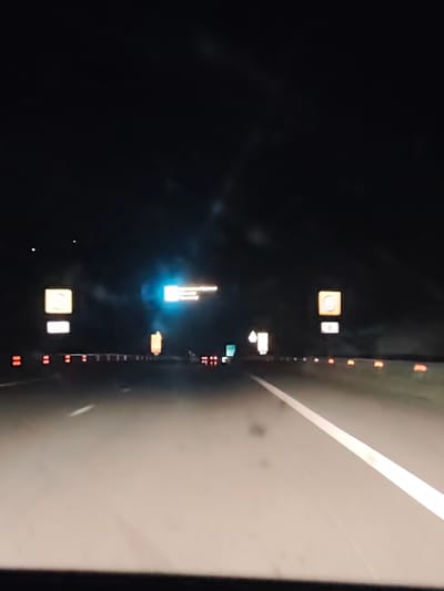 The image depicts a night scene on a highway near Moshtanets, Bulgaria. The road is asphalt with white lane markings. There are roadside reflectors and signs, including one with a speed limit of 80. A blue overhead sign and other traffic signs are visible in the distance. The sky is dark and there are some light sources from other vehicles and signs ahead.