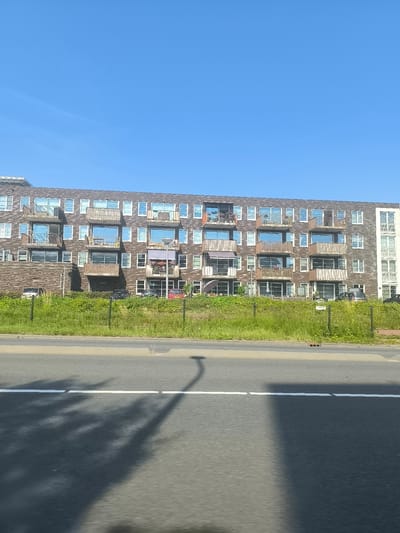 The image shows a modern, multi-story apartment building with brick facade and balconies, located in Aalsmeer, Netherlands. The building has numerous windows and several balconies with wooden railings. In front of the building is a strip of green grass and some vegetation, separated from a paved road by a wire fence. Cars are parked in front of the building. The sky is clear and blue. The foreground shows a paved road with white markings and shadows of trees.