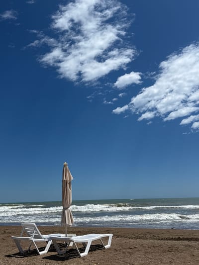 The image depicts a tranquil beach scene on a sunny day. In the foreground, two white plastic lounge chairs and a closed, beige beach umbrella are set up on the sandy shore. The chairs are positioned side-by-side, with the umbrella standing behind them, casting a slight shadow. The beach appears to be composed of dark sand mixed with small pebbles.
The middle ground is dominated by the sea, with gentle waves rolling towards the shore, creating white foam. The water transitions from a dark blue near the horizon to a lighter, greenish-blue closer to the beach.
The background consists of a clear, vibrant blue sky, with scattered white, fluffy clouds occupying the upper portion of the frame. The clouds are wispy and appear to be moving, suggesting a light breeze.
This scene is consistent with a typical beach location within the city of Derbent, Russia, known for its coastline along the Caspian Sea. The image captures a moment of calm and relaxation, with no people present.