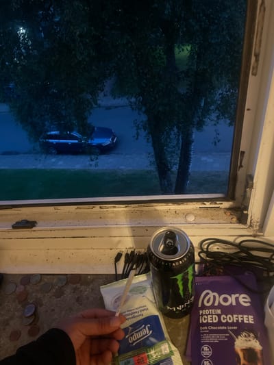 The image shows a cluttered windowsill or surface next to an open window, looking out onto a dark street scene at night or dusk. In the lower right, a person's hand holds a rolled cigarette or joint.
On the surface, an open black Monster Energy drink can with its green logo is prominent. Beside it lies a purple packet of "More Protein Iced Coffee - Dark Chocolate Lover" powder, stating "16g PROTEIN PER SERVING" and "95" (likely calories). A packet of "Jempo Reinigende vochtige doek Hand-Feuchttücher" (wet wipes) is also visible, with text indicating it's "alcohol-free" and "PRODUKT ENTHALT KUNSTSTOFF" (product contains plastic). Several black USB charging cables are coiled near these items. Scattered across the surface are various Euro coins, including 1, 2, 5, 10, 20, and 50 cent pieces. The window frame and the surface itself appear worn and dirty.
Outside the window, dark green trees fill much of the view, with some leaves dimly lit by an unseen light source. A dark-colored car is parked on what appears to be a street or paved area. The dark exterior suggests it is evening or nighttime. The presence of Euro coins and German text on the wet wipes ("Hand-Feuchttücher," "PRODUKT ENTHALT KUNSTSTOFF") indicates a European, likely German, location.