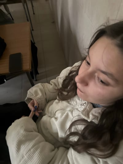 An overhead-angle image depicts a young individual with dark brown, wavy hair, seated indoors in Kalitinovo, Bulgaria. The subject is wearing a light-colored, possibly off-white, corduroy puffer jacket. A metallic zipper is visible on the jacket's upper front. Her left hand, featuring blue nail polish, is visible in the lower-left quadrant of the image. The subject's face is partially shown in profile, looking downwards and to the right, exhibiting no overt expression. To the left of the subject, a light-colored wooden desk or table is present, upon which a dark-colored smartphone lies flat. The background features light-colored tiled walls and a tiled floor, consistent with an institutional or public building interior. Metal furniture legs are discernible beneath the desk. No other individuals are visible.