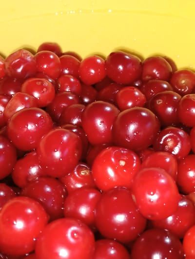A close-up, high-angle shot shows a large quantity of ripe, red cherries filling a yellow container. The cherries are round and plump, with a glossy surface reflecting the light. Some of the cherries have small water droplets on them. The background is a solid, bright yellow. The image does not contain any elements that would indicate a specific location in the city of Баяндай, Kazakhstan, or any people, actions, or interactions.